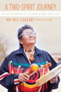 Book cover for A Two-Spirit Journey. Ma-Nee Chacaby, an Oji-Cree person, is gazing up with a peaceful expression and holding a drum and rattle. Ma-Chee is wearing a black ribbon-shirt with blue, red and yellow ribbons.